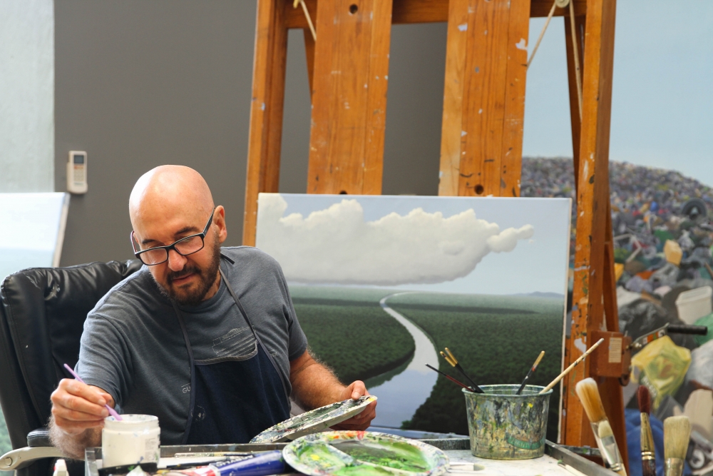 Color photographic portrait of Tomás Sánchez painting a river scene in his studio