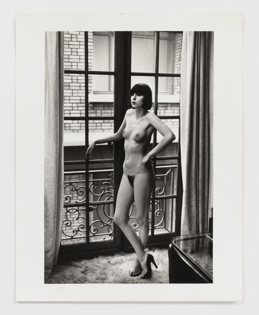 Black and white photographic print by Helmut Newton of a nude woman standing in front of a wndow