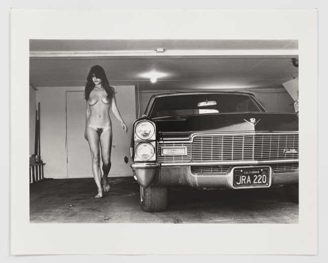 Black and white photographic print of a nude woman standing beside a car