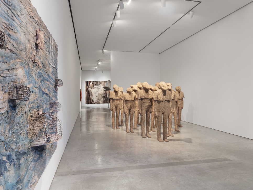Installation view of large scale textured canvas with birdcages by Anselm Kiefer and group of multiple burlap and resin life-size standing figures with different heads by Magdalena Abakanowicz along with her textiles and a steel bird sculpture