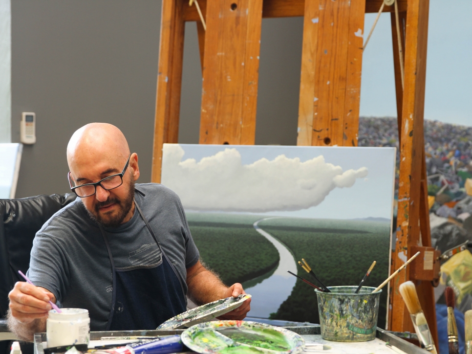 Color photographic portrait of Tomás Sánchez painting a river scene in his studio
